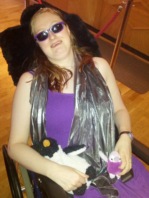 Danni at the prom. They are wearing a purple dress, a silver stole, sunglasses and a tiara. They are holding two penguins, a larger black and white one called Penguin, and a smaller sparkly purple one called Purple Penguin.