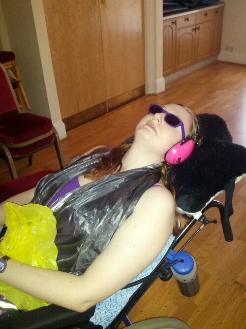 Danni lying in their wheelchair at the Prom. They are wearing sunglasses and pink ear defenders, and holding a sick bag.