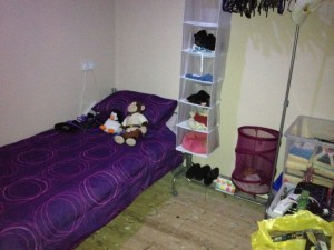 A small bedroom with a single bed in the corner with purple bedding, a clothes rail with hanging shelves, and boxes with items in. There is clear floor space in the middle.