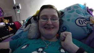 Danni is lying in bed with a massive smile on their face. On their shoulder there is Poseidon, a baby emperor penguin.