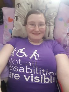 Danni is in bed, and has clean, tidy short hair. They are smiling, and wearing a purple t-shirt saying 