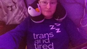 Danni is lying in bed wearing a purple top saying "trans and tired" with "zzz" above. They look tired.
