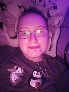 Danni is lying in bed with a small smile. The image is tinted purple from the light. 