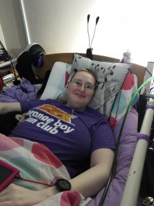 Danni is smiling happily at the camera in bed. They are wearing a purple Orange Boy Fan Club top with an orange cat on it.