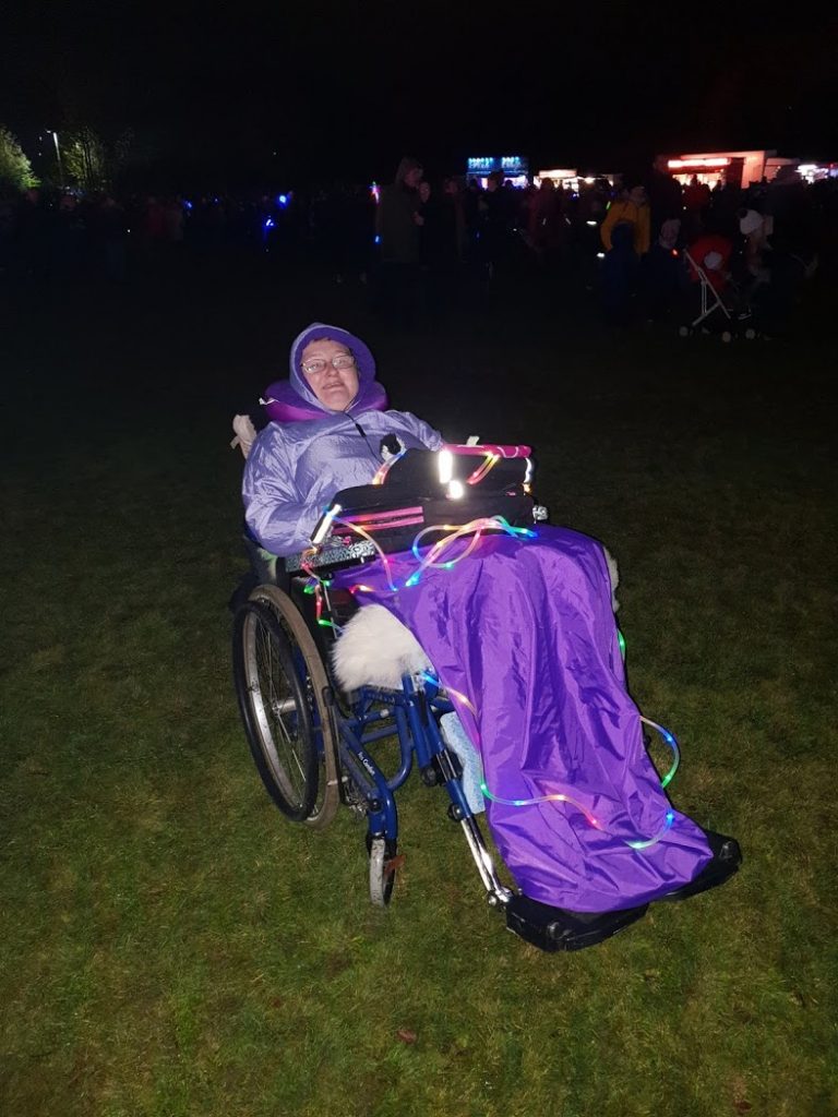 Danni at the local fireworks event. They are in their wheelchair, wearing purple waterproofs, with a tablet on their lap and a light rope around their body.