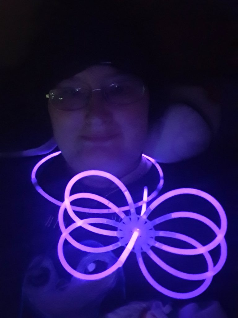 A very dark photo of Danni. Their face can just be seen by the light of a purple glow stick necklace and glow stick toy. 