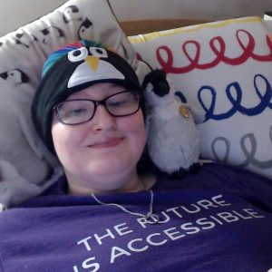Danni is lying in bed, on a penguin cushion. They have very pale white skin and blue eyes. They are wearing purple glasses, a rainbow turban, a penguin headband-type headphones, a purple top saying "The future is accessible"in whire writing and a silver necklace with a wedding ring. They have a grey baby emperor penguin on their shoulder and are smiling.