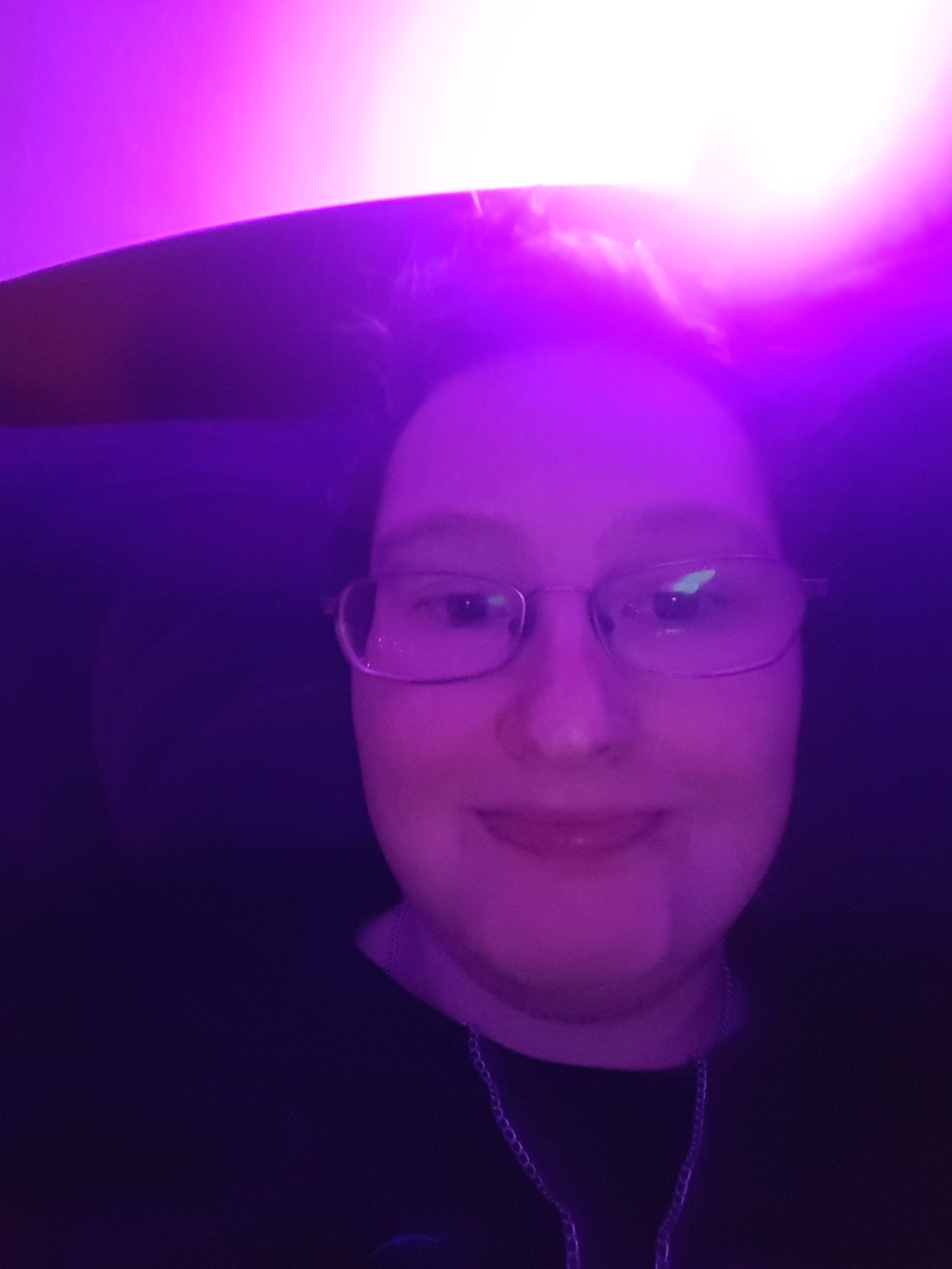 A photo of Danni's face. Their head is resting on a pillow. The entire image is tinted purple from the light in the top right corner.