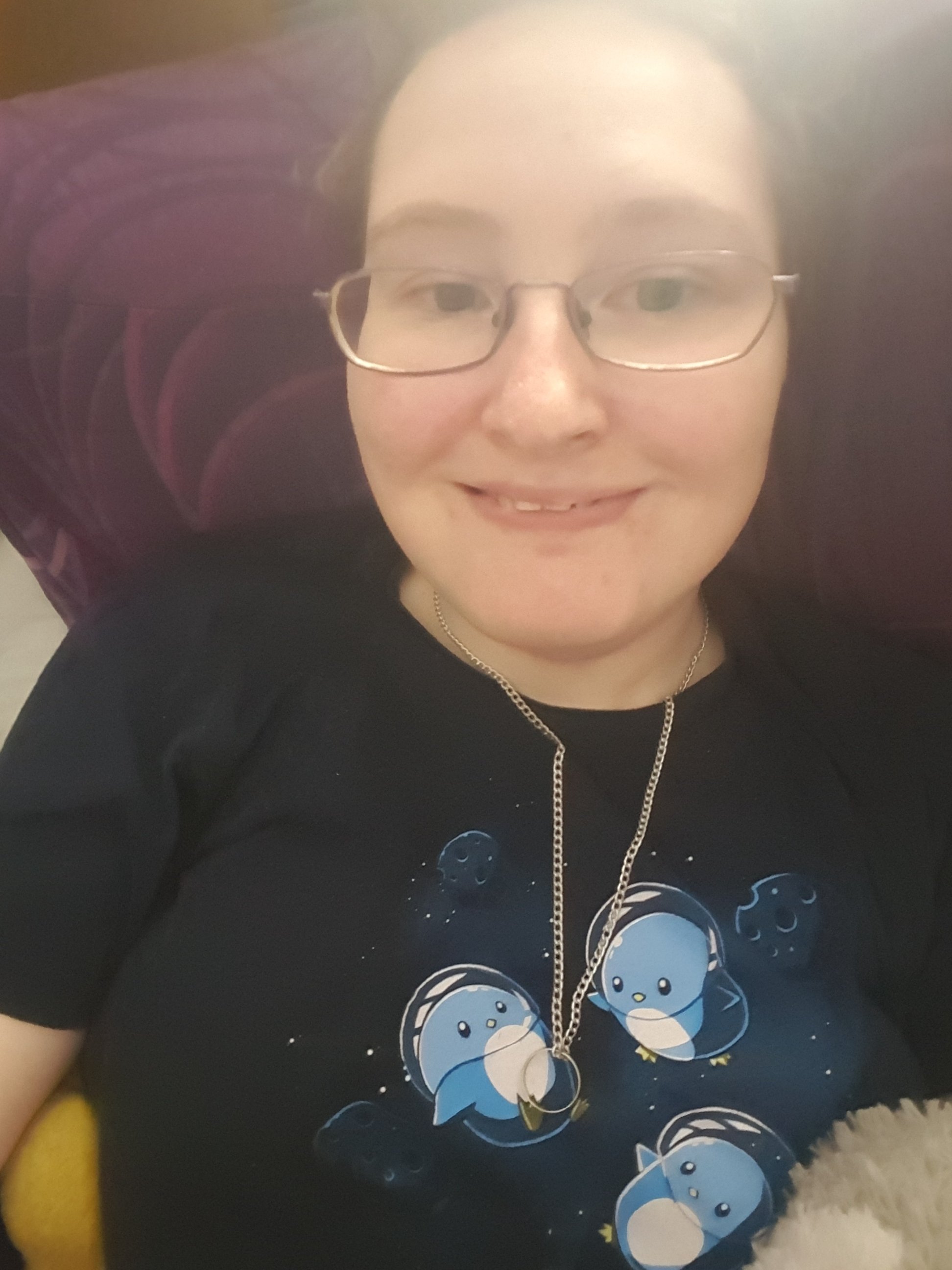 A photo of Danni lying in bed, wearing a dark t-shirt with blue penguins wearing space helmets. They are smiling. 