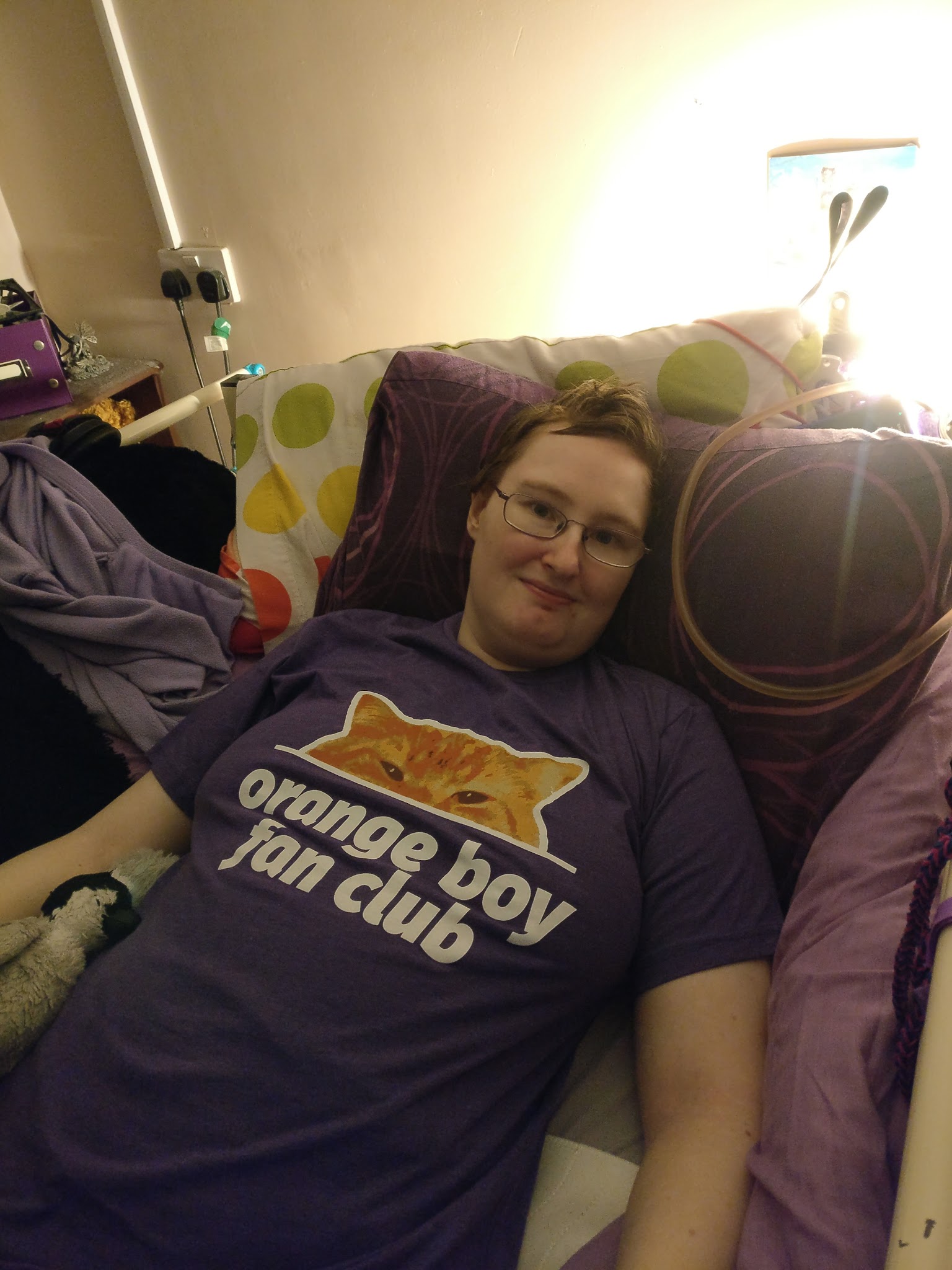 Danni is lying in bed, wearing a purple t-shirt with a picture of the top half of an orange cat's face (Bilbo), with orange boy fan club written underneath in white. 