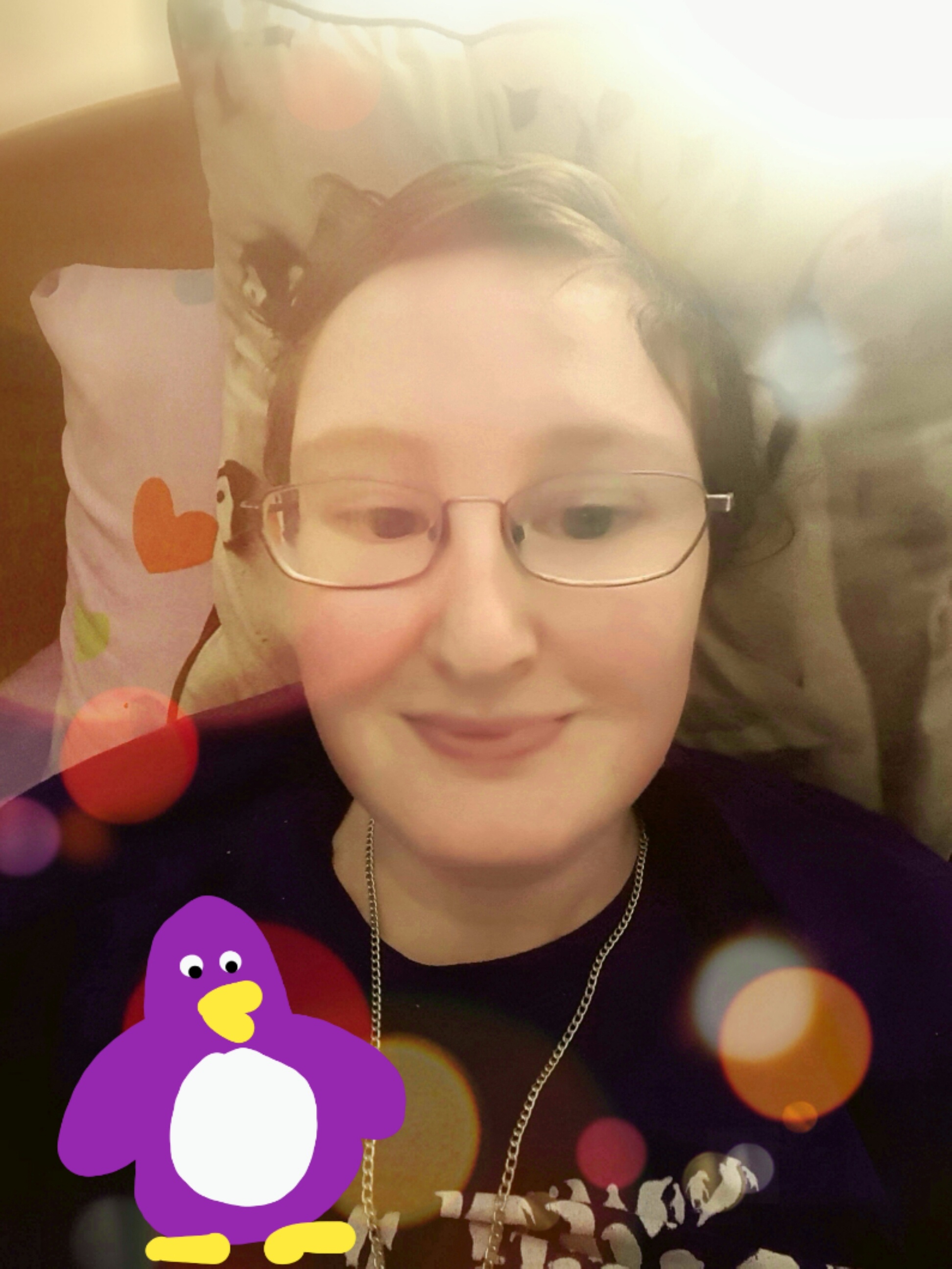 Danni is lying in bed. The image has been edited with various filters, making it look smooth, have bigger eyes, has colourful circles around their head, and has a hand drawn purple penguin in the bottom left corner. 