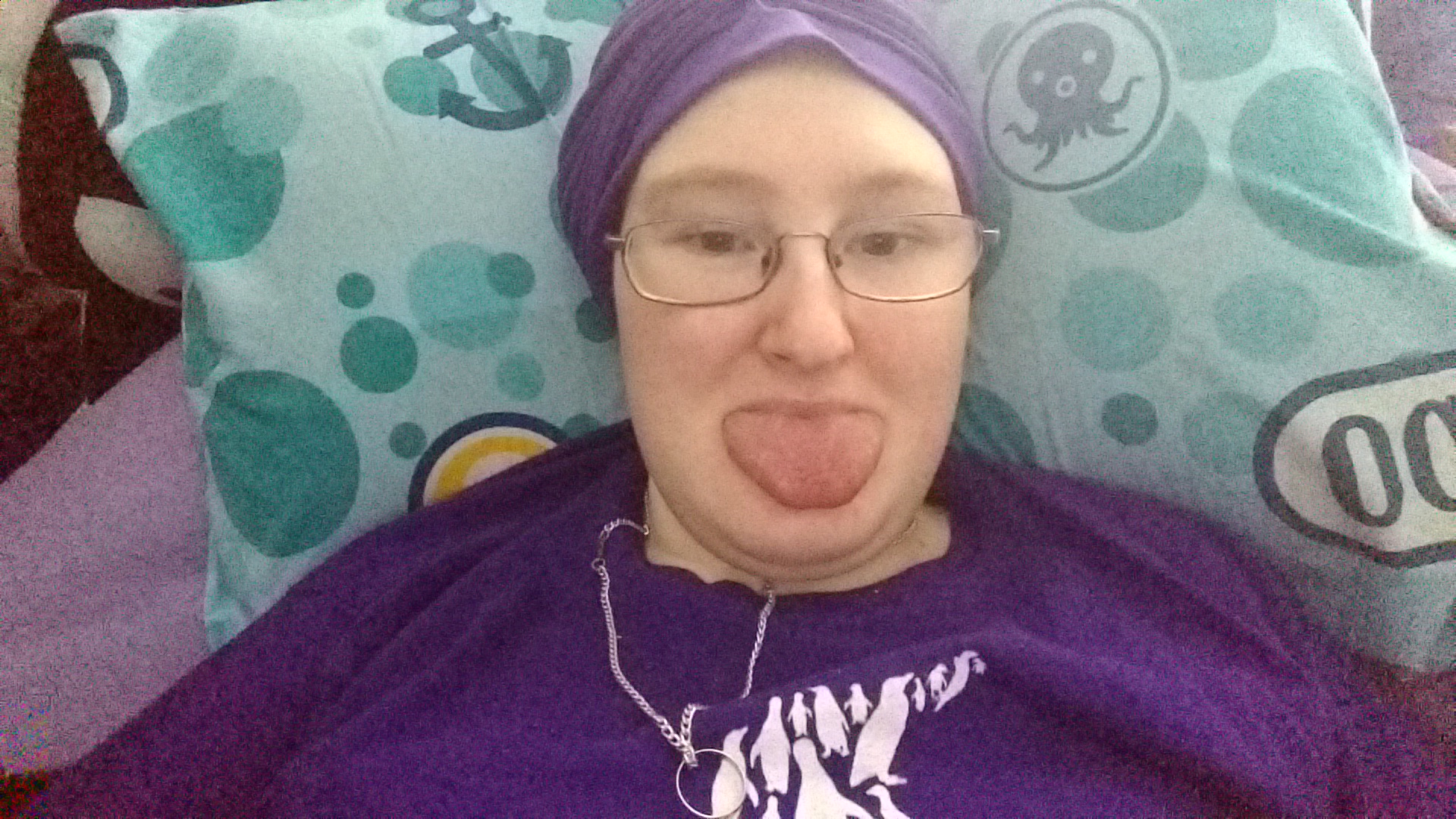 Danni is lying in bed wearing a purple turban. They are sticking their tongue out. 
