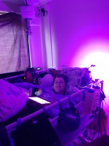 Danni is lying in bed in their back. They are smiling. The image is tinted purple from a light behind the bed. 