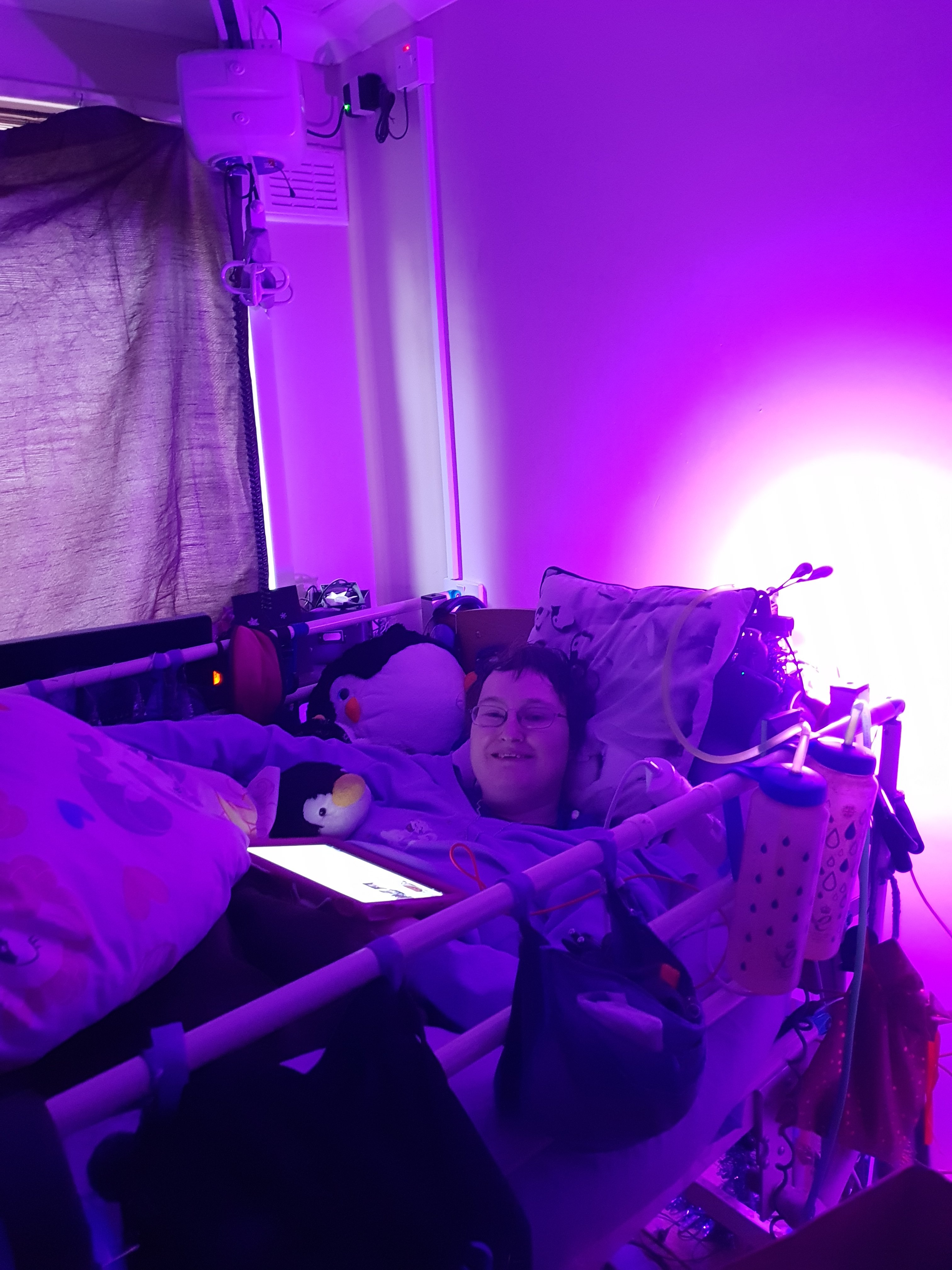 Danni is lying in bed in their back. They are smiling. The image is tinted purple from a light behind the bed. 