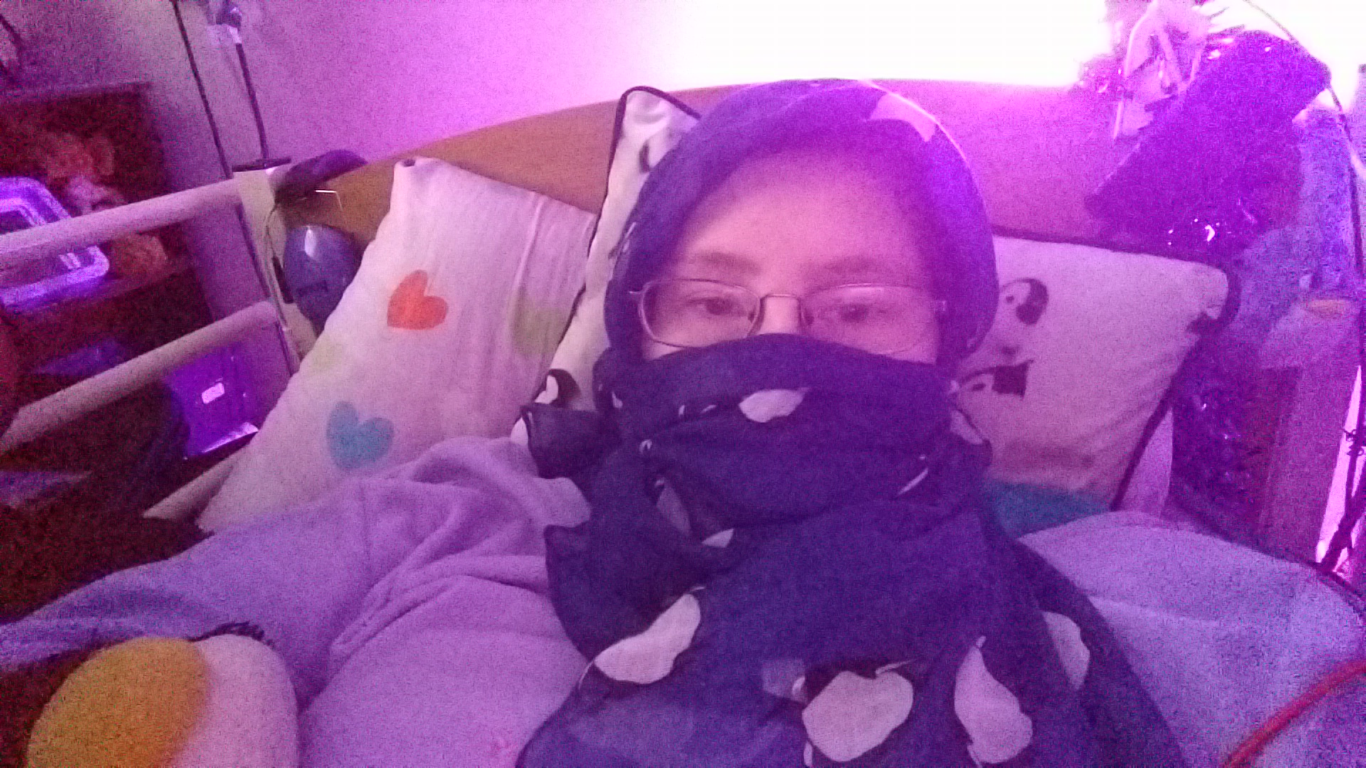 Danni is lying in bed, with a dark blue scarf with penguins on wrapped around their head and lower face. The photo is tinged purple from the light. 