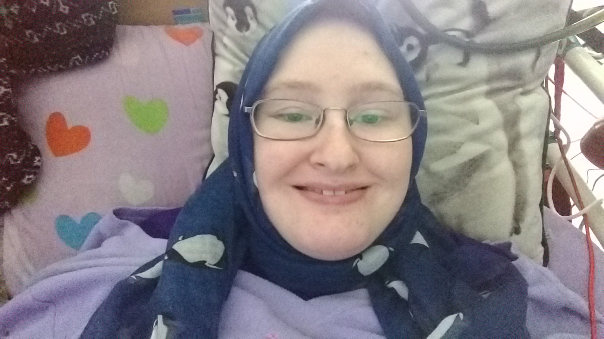 Danni is lying in bed, wearing a blue penguin scarf over their hair. They are smiling. 