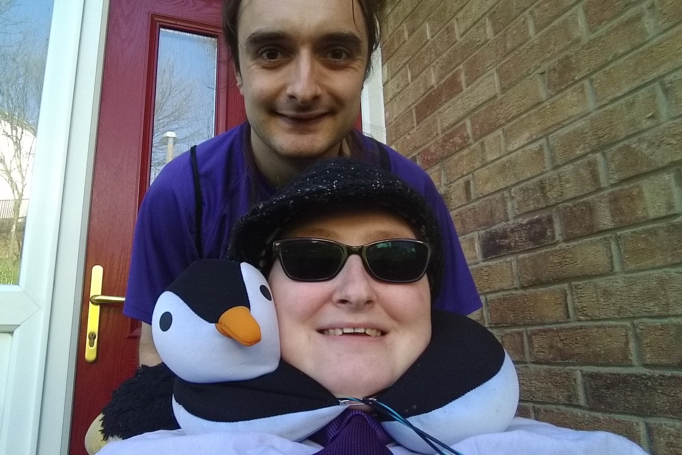 Danni is at the bottom of the image, wearing a shirt and tie, sunglasses and a hat. Above is Johan, who is wearing a purple t-shirt and is leaning down. They both are smiling and are outside. 
