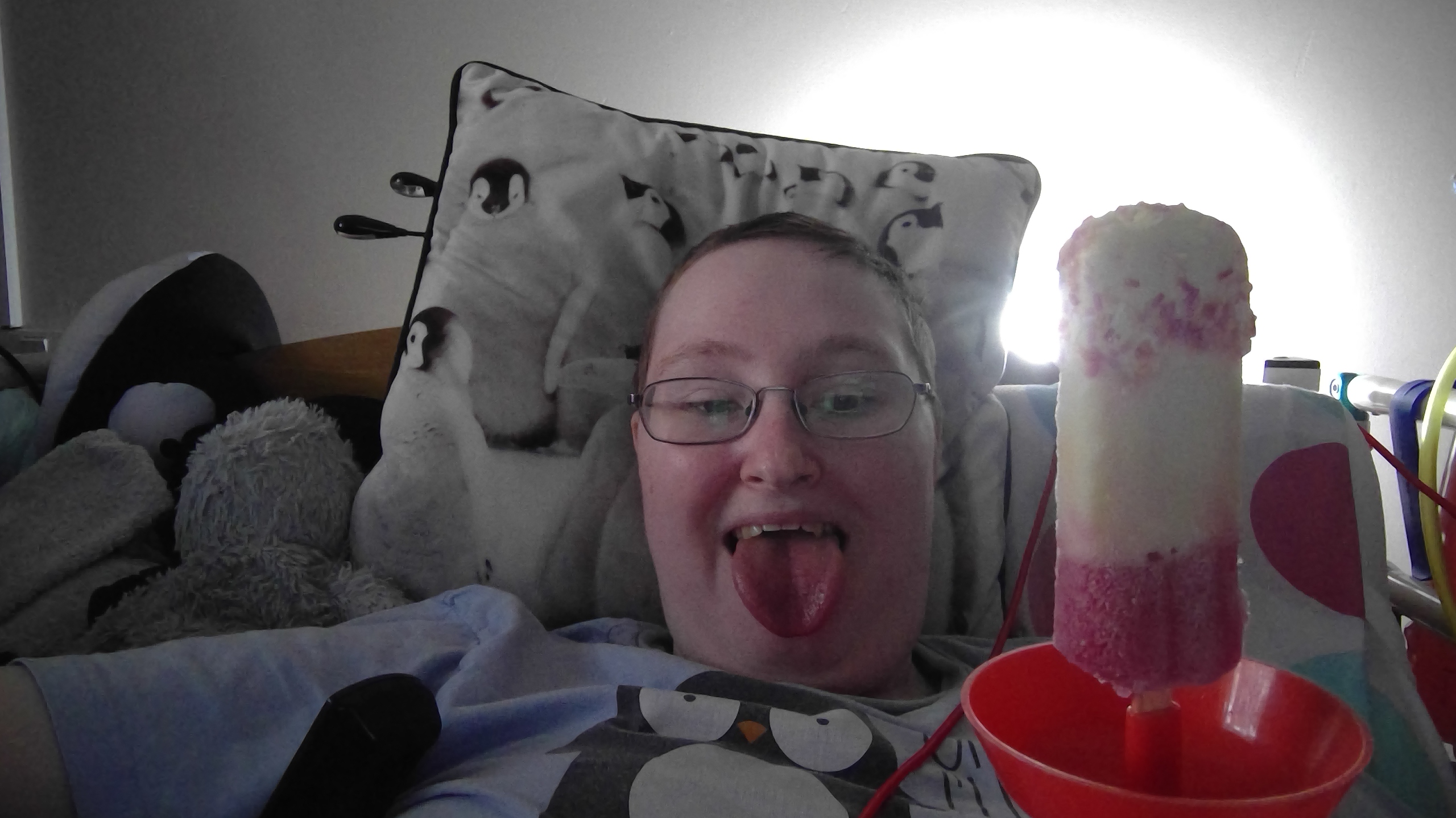 Danni is lying in bed, sticking their tongue out while holding a Fab ice lolly in a red holder. 