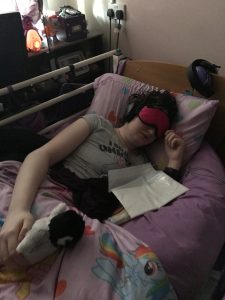 Danni is lying in bed. They have an eye mask on that also has ear muffs, a sick bag in front of them, and their left hand is resting on a penguin.