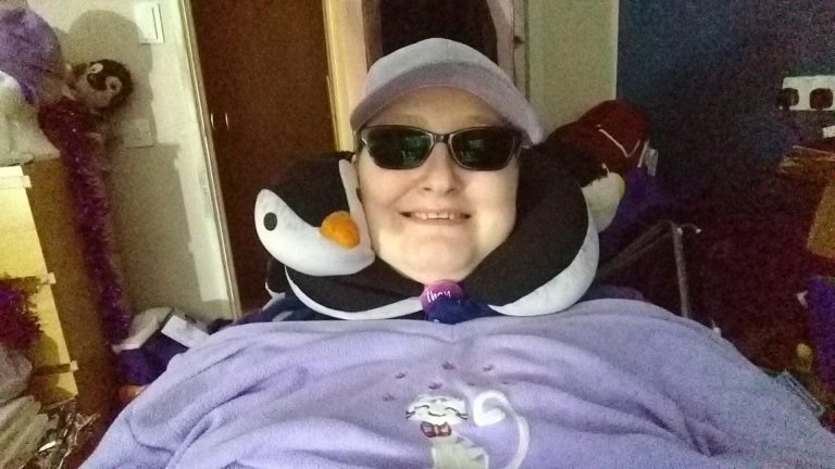 Danni is in their wheelchair, wearing a lilac top with a cat on it, a lilac baseball cap, sunglasses and a penguin neck cushion. They are smiling.