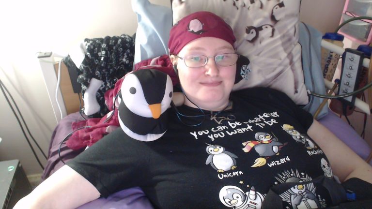 Happy Danni in bed, wearing a maroon headscarf with penguins on, and a black t-shirt with 6 penguins, showing various things you can be. 