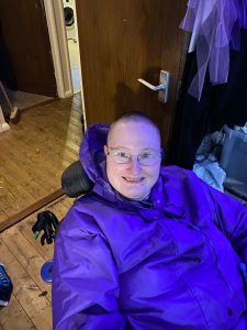 Danni in their wheelchair. They are smiling and have a shaved head with only a tiny amount of hair left, and are wearing a purple coat and glasses. 