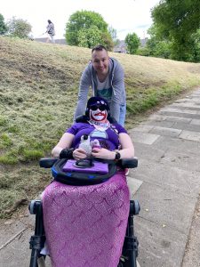 Danni and Martin outside. Danni is in their wheelchair, wearing a purple hat, purple sunglasses, a Pizza John mask (man's face in red and white with a moustache), a purple penguin top, pink bib and purple mermaid tail. They are holding Poseidon (a baby emperor penguin) and Purple Penguin.

Martin is standing behind, leaning on the wheelchair. He is a tall white man with short brown hair, a big smile, and is wearing a grey hoodie, white t-shirt with blue and green image, and blue jeans.