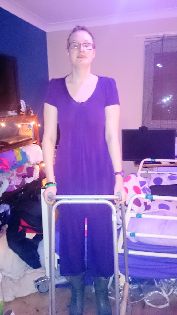 Danni, a tall white person with light brown hair and glasses, standing with a zimmer frame in front of a bed. 

They are wearing a purple nightdress, purple pyjama bottoms and green heeled boots.
